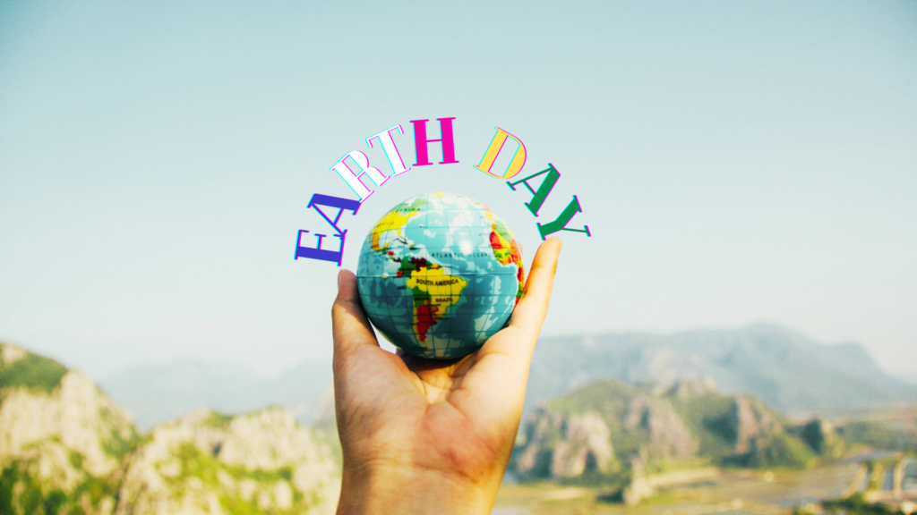 A hand holding a small globe with "earth day" text overlaid, symbolizing environmental awareness against a mountainous backdrop.