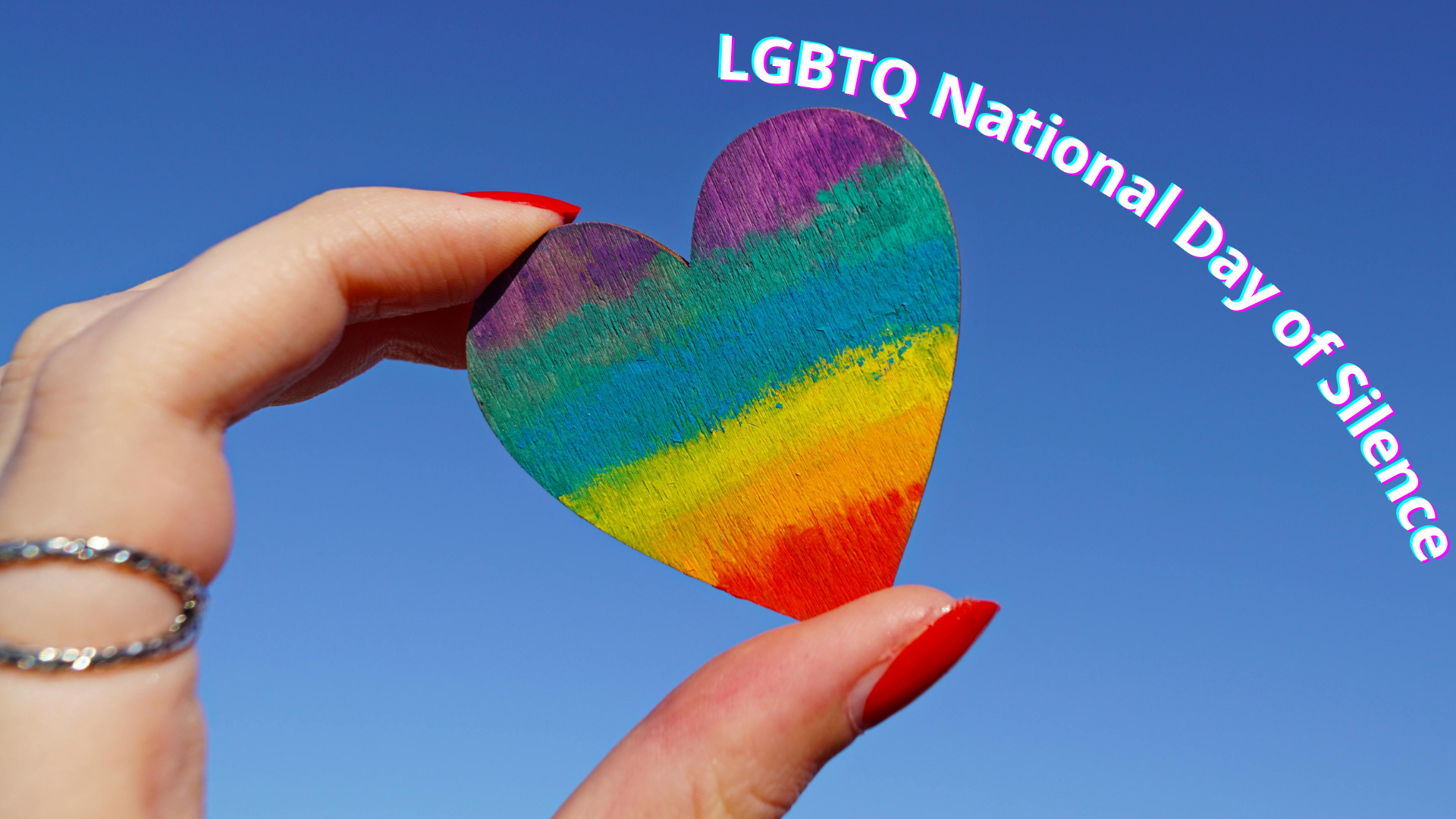 A hand holding a rainbow-painted heart cutout against a blue sky, symbolizing support for lgbtq national day of silence.