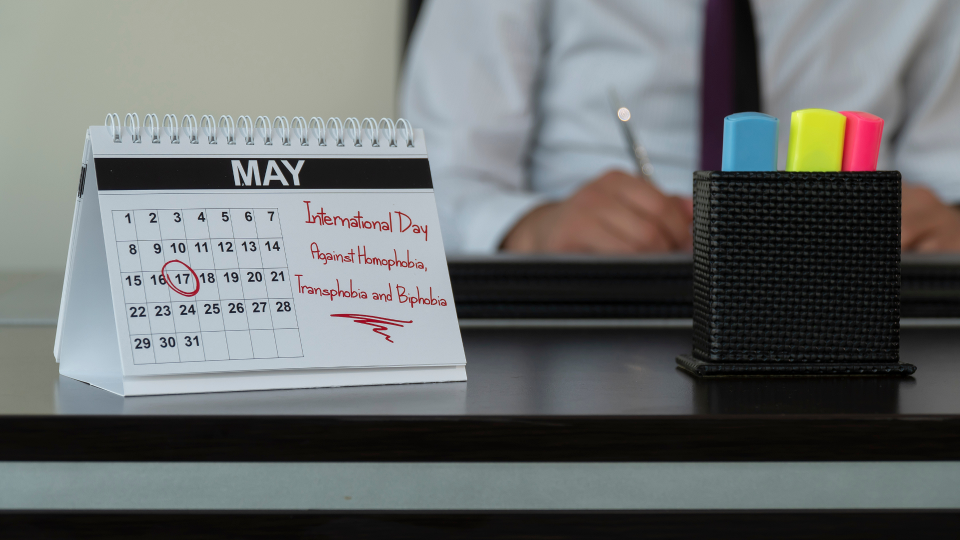 Desk calendar showing "international day against homophobia, transphobia and biphobia" on may 17, with a person working in the background.