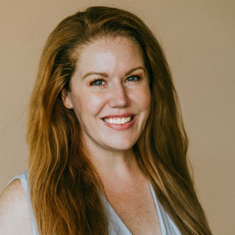 A person with long, reddish-brown hair smiles while posing against a plain beige background.