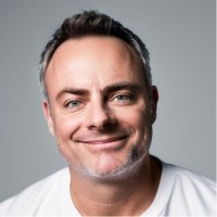 Smiling man with short hair and a light beard, wearing a white shirt, against a plain background.