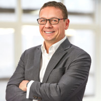 A man wearing glasses and a gray suit jacket over a white shirt stands with his arms crossed, smiling, in front of a blurred indoor background.