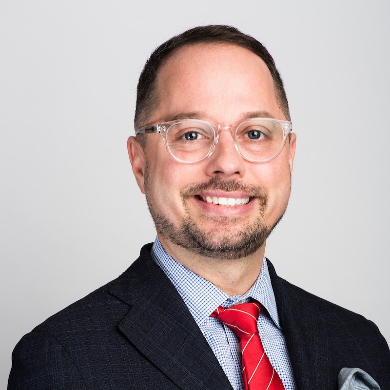 Man with short hair, beard, and glasses wearing a dark suit, light blue shirt, and red tie, smiling against a plain white background.