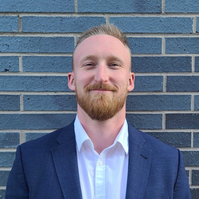 A man with a beard and mustache, wearing a white shirt and navy blue blazer, smiles while standing in front of a gray brick wall.