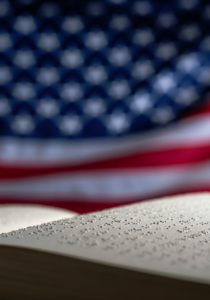 A Braille page is in focus in the foreground, with a blurred American flag in the background.